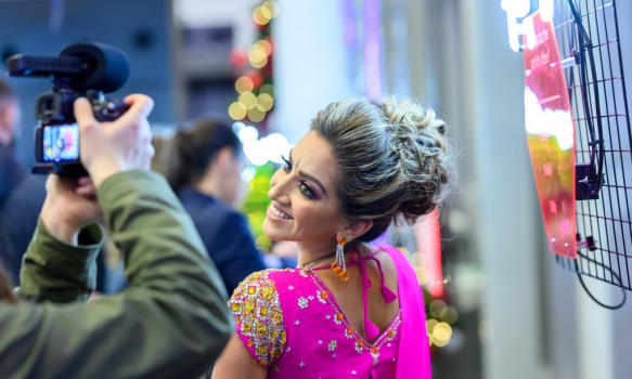 Image shows Ria Meera Munshi posing for a photograph in front of a camera wearing pink.