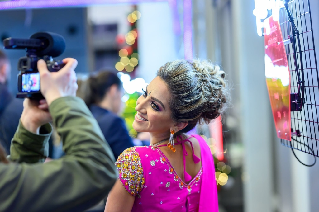 Image shows Ria Meera Munshi posing for a photograph in front of a camera wearing pink.