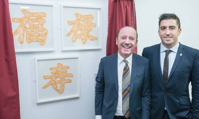 This image shows two men. Patrick Trodden a chef from The University of Salford. and Stephen Lowy a former student. Patrick is wearing a navu blue suit, white shirt and striped orange tie. He has brown hair around the back and sides of his head and is smiling. Stephen Lowy wearing a navy blue suit, blue shirt and black tie whilst smiling. He has dark quiffed hair.