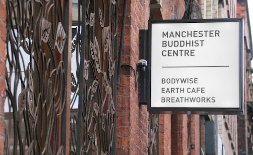 This photo shows the side of the Manchester Buddhist Centre's exterior, which has a white square sign coming off the wall that says 'Manchester Buddhist Centre. Bodywise, Earth Cafe, Breathworks'. 