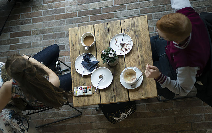 Two students sat at a wooden table in the GK Gallery and Cafe, Salford. On the table there is two plates with scrunched up napkins and crumbs on. There is also two coffee mugs, saucers and a lovely potted plant as a centre piece. 