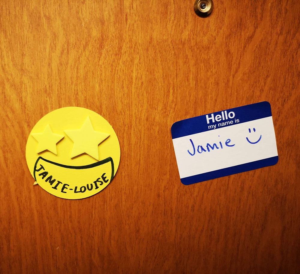 Stickers on a wooden door, one is in a yellow circle shape with stars for eyes and the name 'Jamie-Louise' penned on. The other sticker is a typical 'Hello my name is' sticker with 'Jamie' and a smiley face drawn in the white center.