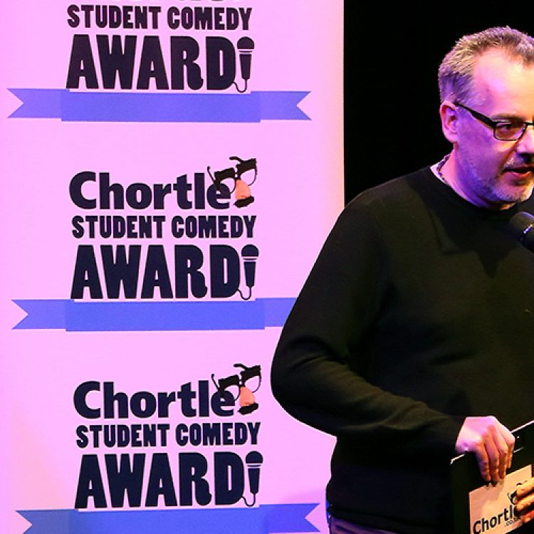 Steve Bennett, manager of chortle hosting on the main stage in front of two flats with the 'Chortle Student Comedy Awards' logos on