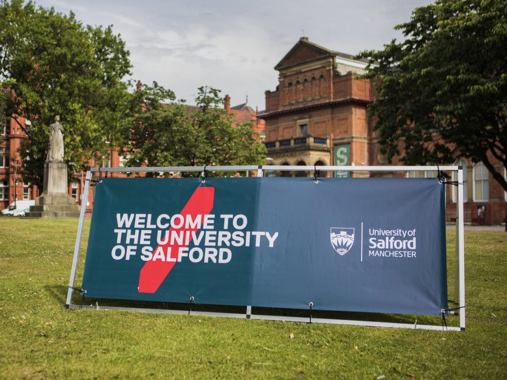 In the background is the Salford Museum and Art Gallery as well as some fully bloomed trees, a statue and green grass. In the foreground is a horizontal sign with our logo on and the words 'Welcome to The University of Salford'.