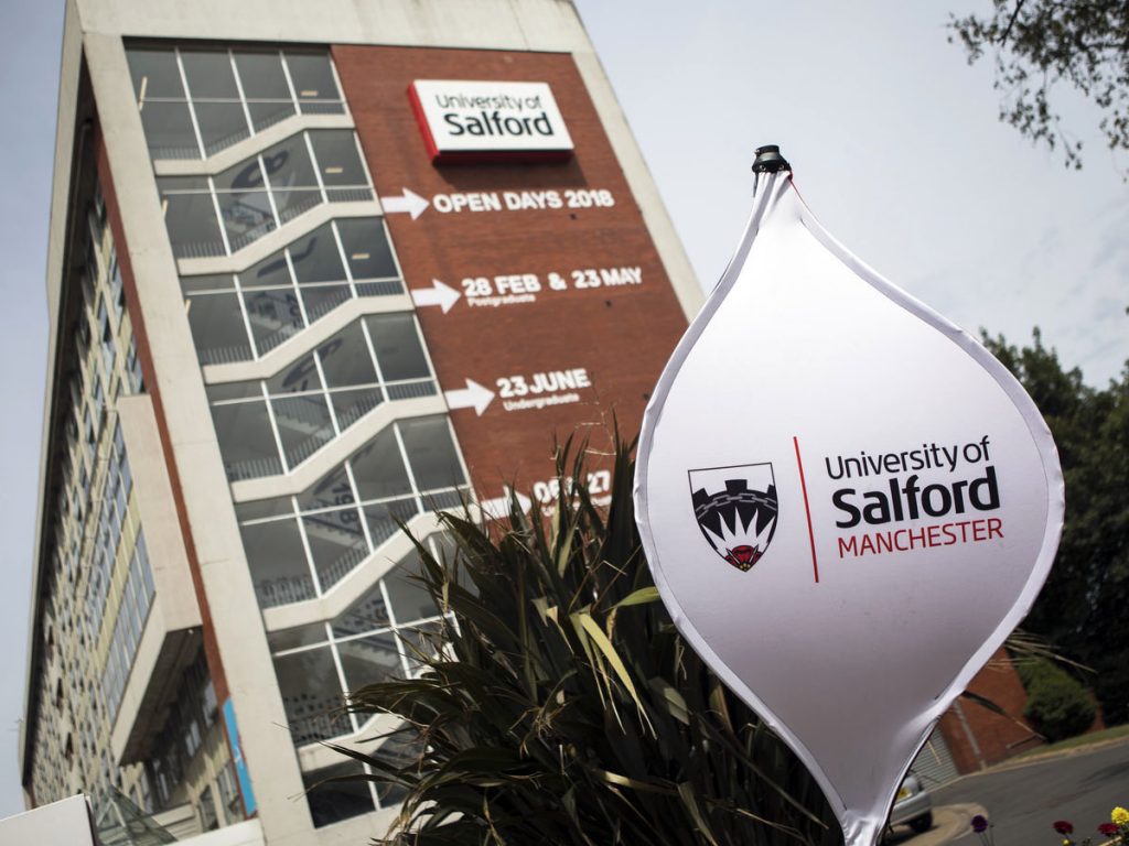 In the background is the Maxwell Building with each 2018 open day written alongside the windows. In the foreground is a plant and a spinning sign that reads 'University of Salford Manchester'