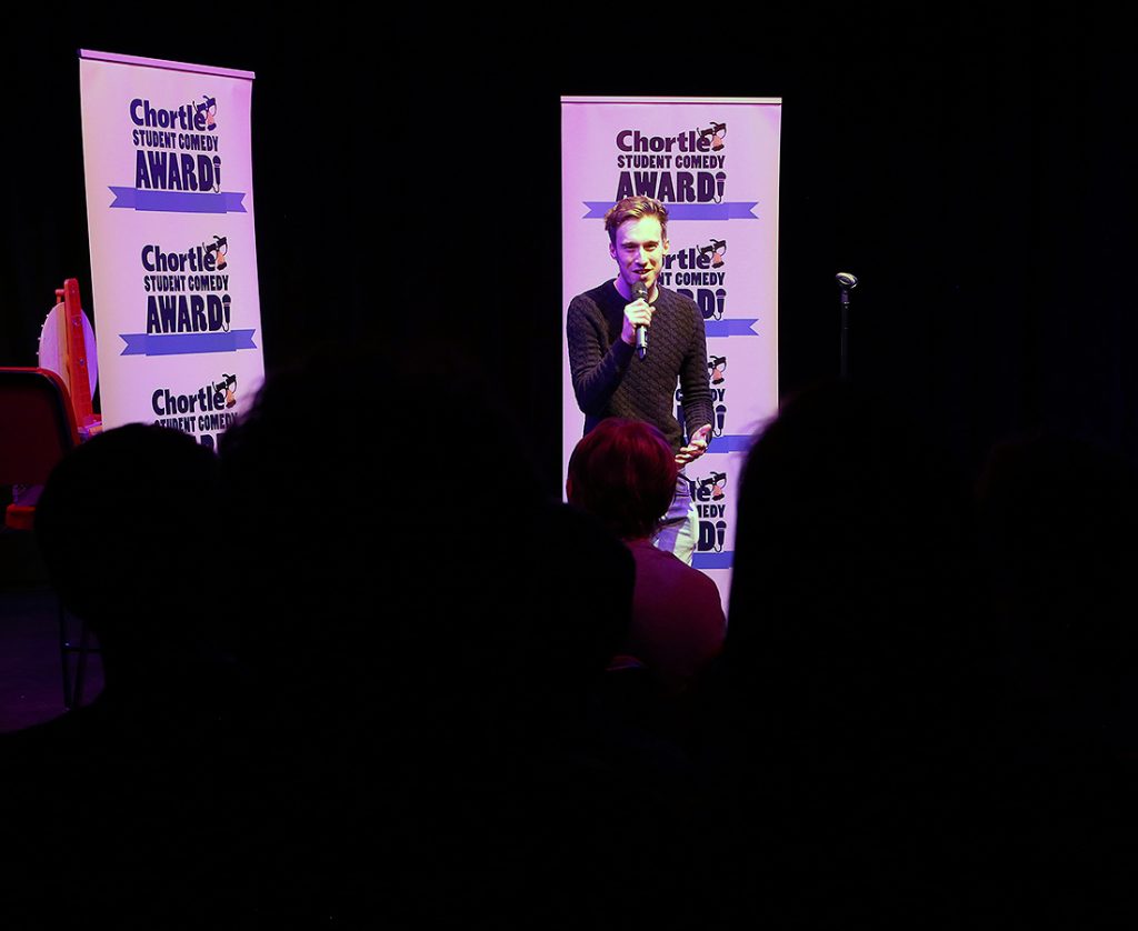 Callad Gale wearing a dark grey jumper performing in front of a big crowd. He is stood in front of two flats with the Chortle Student Comedy Award logos on it.