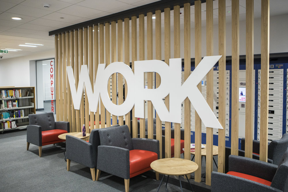 From the recently revamped Clifford Whitworth Library, this image shows some of the shelves and seating arrangements of the ground floor as well as a wooden panel divider that has the words 'WORK' on it in white.