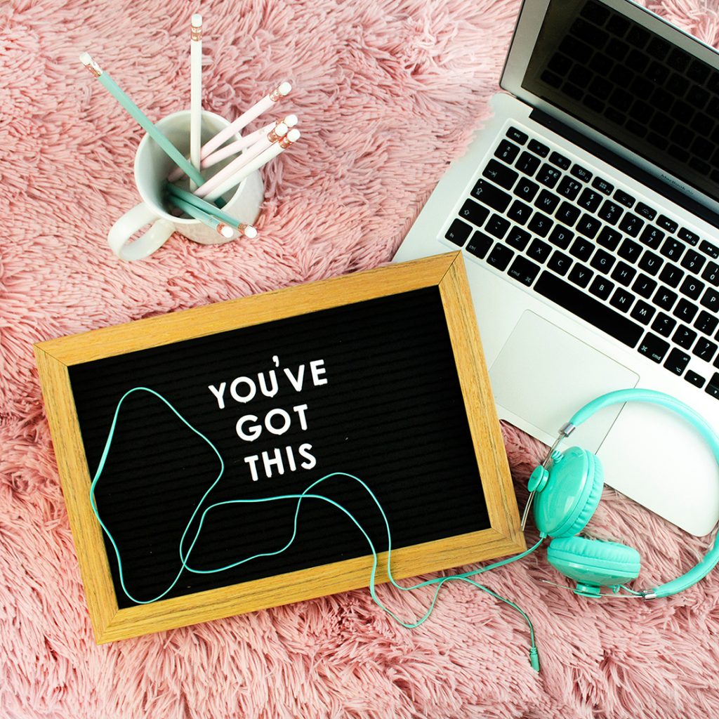 Laid on a pink fluffy carpet, there is a mug full of turquoise and white coloured pencils, a laptop, a pair of green headphones and a custom peg board that has the saying 'You've got this' on it.