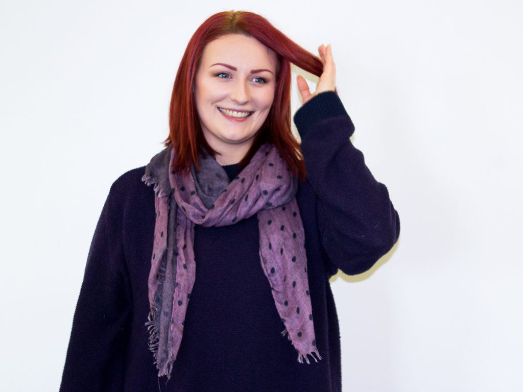 Photo of Hazel, who is wearing a purple jumper and a lighter purple scarf stood in front of a white wall. She is smiling at the camera with one hand fiddling with her hair.