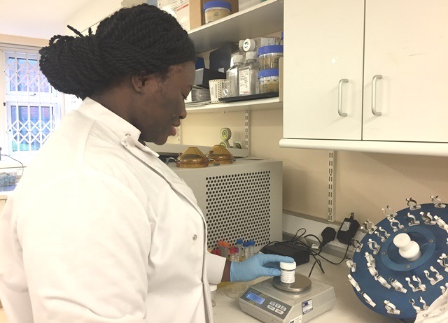 Suzan is stood in a white lab coat in front of a counter-top full of science equipment.