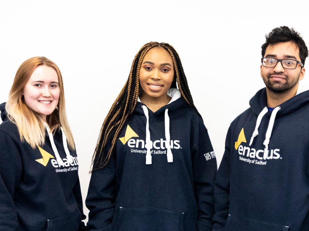 Three other students involved in Enactus. All three of them having the navy Enactus logo hoodies on. They are stood facing the camera and smiling.