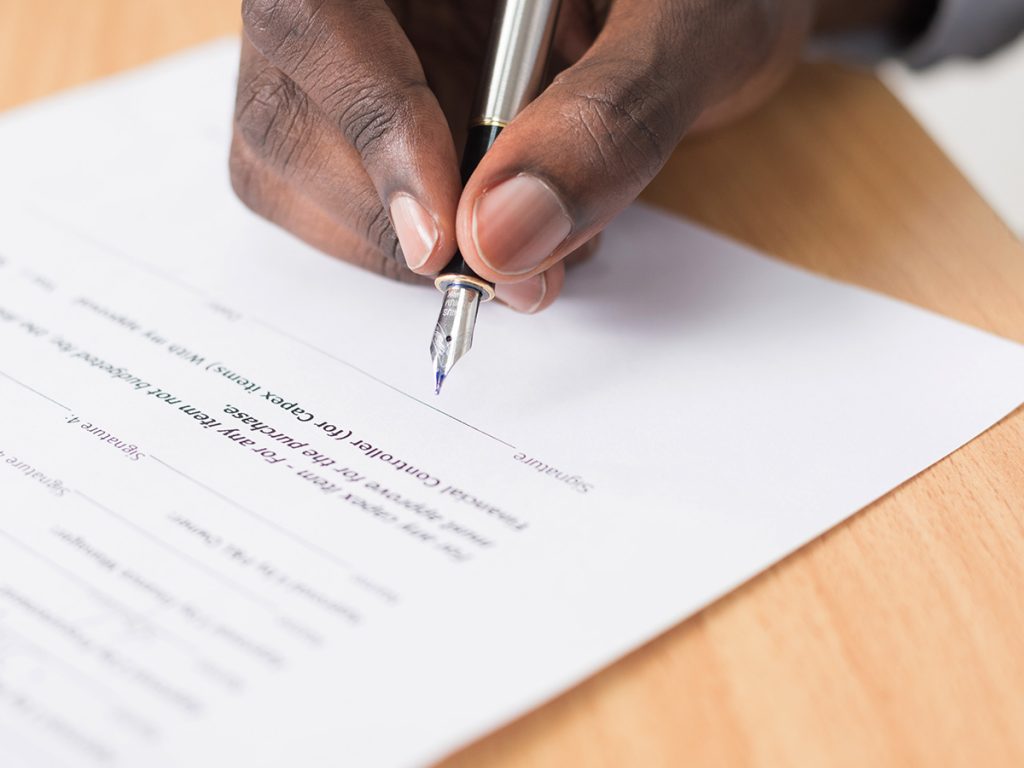 A man is using a fountain ink pen to sign a contract or document of some form which sits on top of a wooden table.