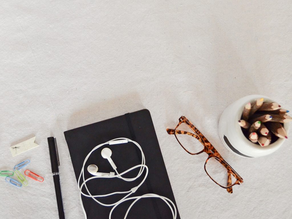 A black notebook sits on a white table with white headphones tangled on top. On the left next to it, is a black pen and some assorted colour paper clips. On the right of the notebook is a pair of glasses and a pot full of sharpened coloured pencils.