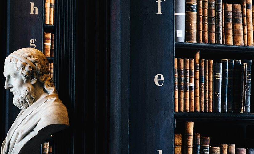 A dark black wood bookcase with tons of books and a ladder, in the distance you can see marble busts of different male figures