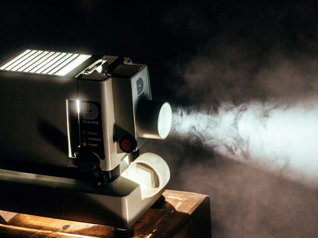 A Dukane projector sits on a wooden table. It is turned on projecting light forward. 