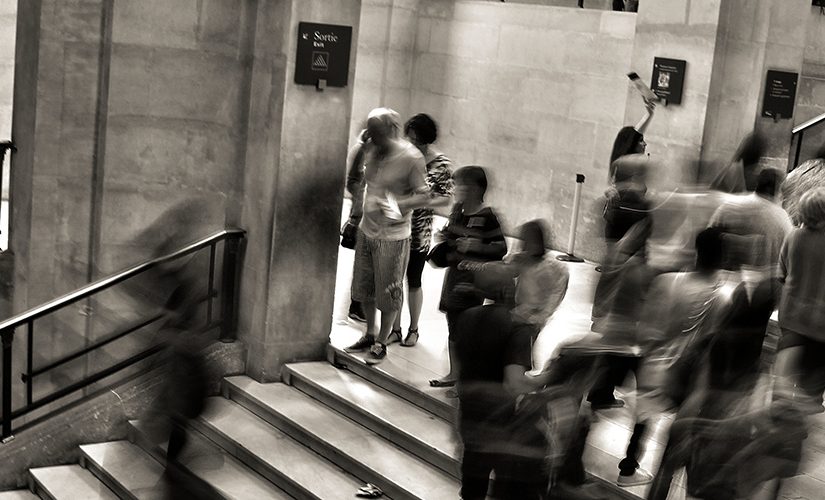 Edited in dark sepia/black and white this is a photo taken with adjusted shutter speed. It shows people and crowds moving throughout a station, people's movement is blurred.