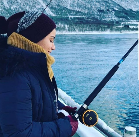 A photo of Marwa wearing a striped knitted beanie, a yellow scarf and navy coat with a fishing rod looking out to a cold looking lake.