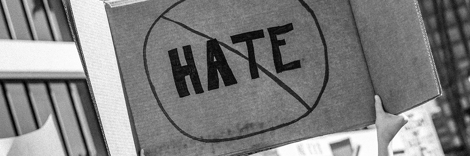 Taken or edited in black and white, someone is holding up a cardboard protest sign that shows the word 'HATE' crossed out to represent being against hate crimes