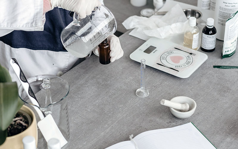 On a grey wooden desk someone has set up a workspace that consists of digital scales, glass laboratory equipment, mortar and pestle and a notebook. The person wearing medical disposable gloves and a lab coat is pouring something from a flask into a bottle.