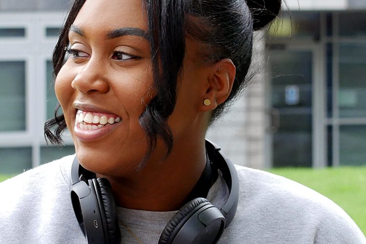 Artiana is sat in front of the New Adelphi building wearing a grey jumper and black headphones around her neck. She is looking into the distance and smiling.