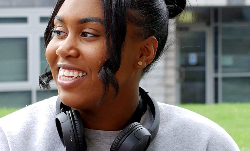 Artiana is sat in front of the New Adelphi building wearing a grey jumper and black headphones around her neck. She is looking into the distance and smiling.