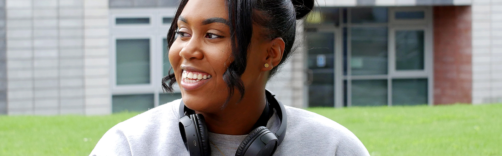 Artiana is sat in front of the New Adelphi building wearing a grey jumper and black headphones around her neck. She is looking into the distance and smiling.