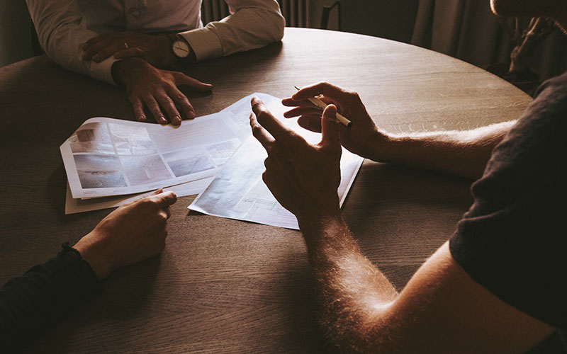 A group of people (faces not shown) gathered around a round wooden table with sheets of papers in front of them. It looks like a semi-formal meeting due to the team wearing formal shirts and watches. 