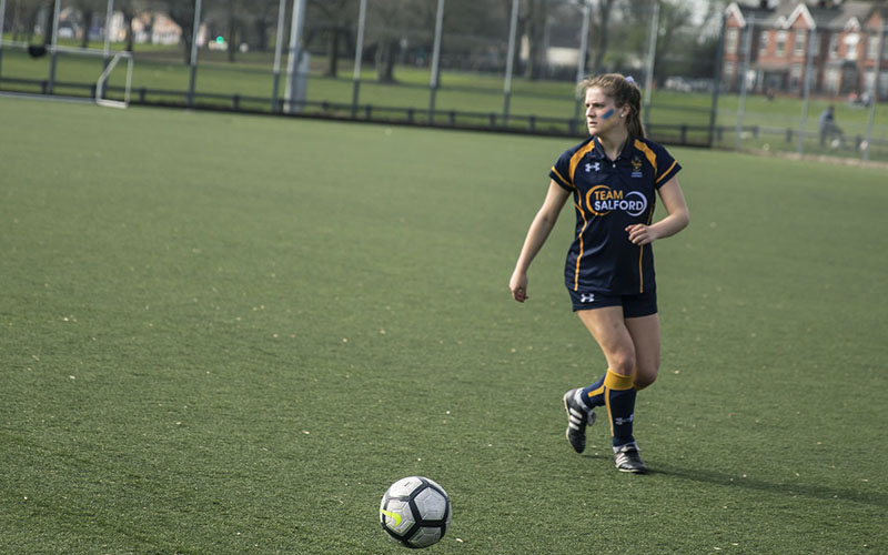 A photo of a girl playing football taken on a green grassy pitch in the Spring. The weather is nice and looks sunny. The girl is wearing a 'Team Salford' football kit which consists of colours navy blue and yellow. She also has blue and yellow face paint stripes on her cheeks. She looks like she is gearing up to take a kick of the football.