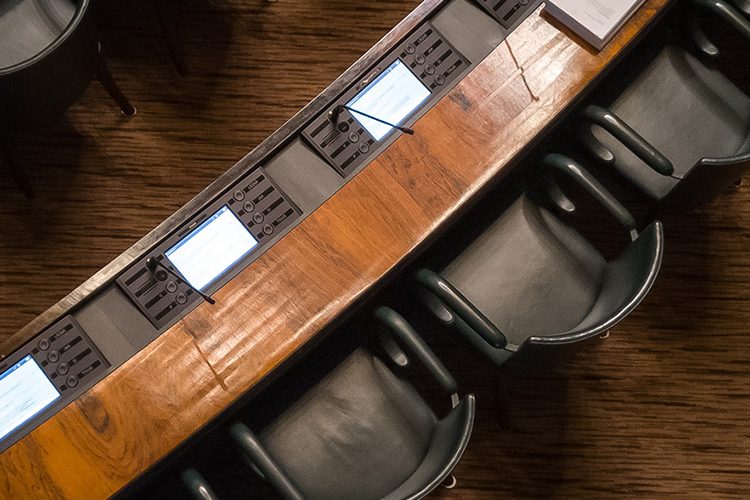 An up-top shot of several black armchairs in front of wooden desks, set out like a meeting room or parliamentary discussion.