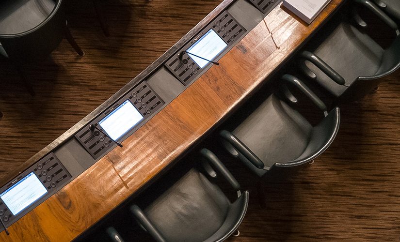 An up-top shot of several black armchairs in front of wooden desks, set out like a meeting room or parliamentary discussion.