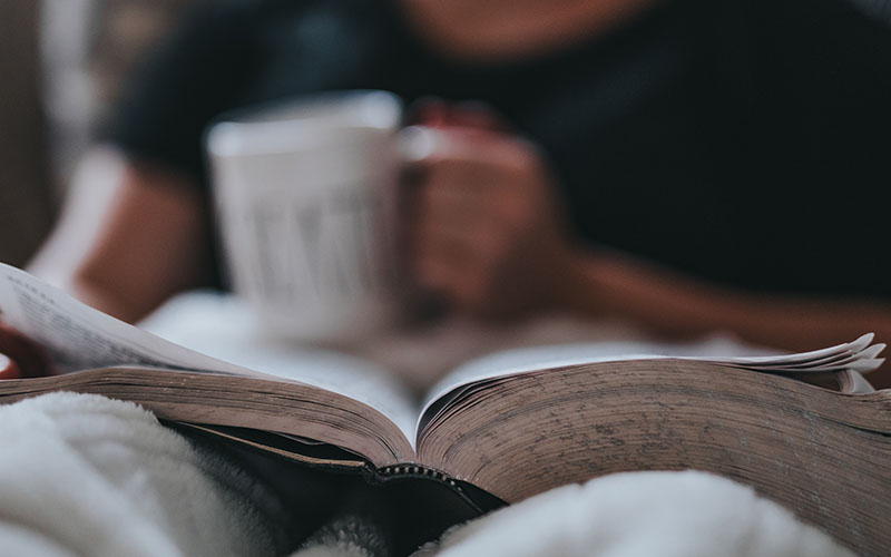 A close up shot of someone lying underneath a blanket with an open book on their lap holding a white mug. 