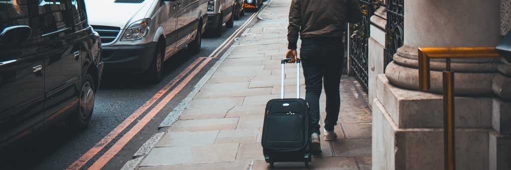 A photo of a person walking down a street pulling a black wheeled suitcase behind him. 