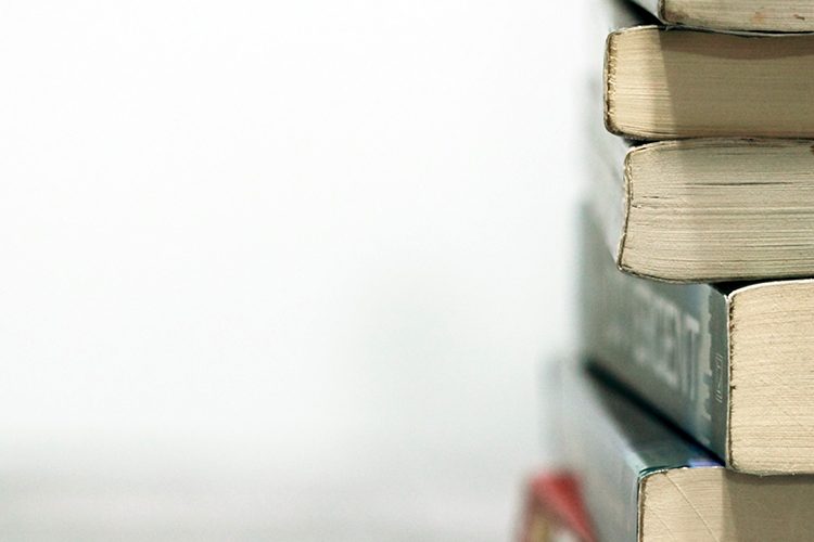 On a white background, a stack of books sits on a desk, the bottom ends of the books facing the camera.