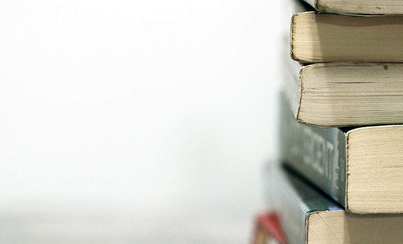 On a white background, a stack of books sits on a desk, the bottom ends of the books facing the camera.