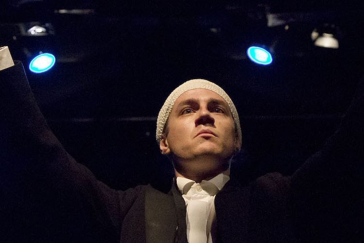 Performer Ryan O'Shea stands at the front centre of the stage, with a bandage wrapped around his scalp. He is holding a baton up as if to conduct an orchestra.