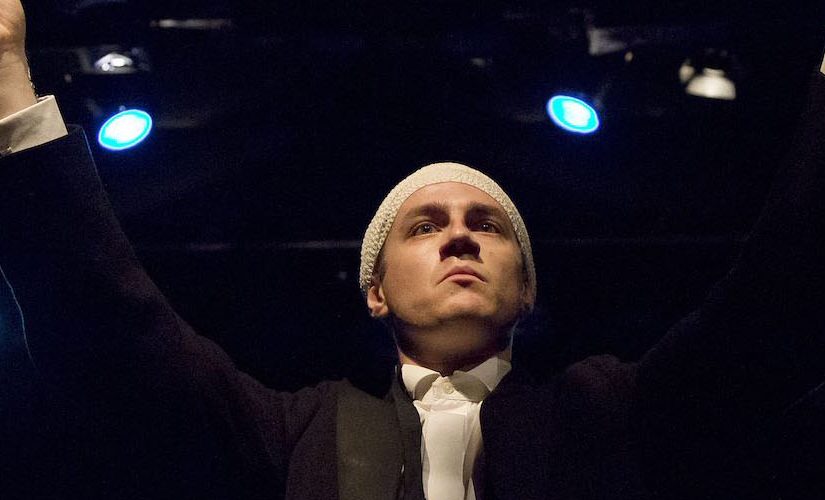 Performer Ryan O'Shea stands at the front centre of the stage, with a bandage wrapped around his scalp. He is holding a baton up as if to conduct an orchestra.