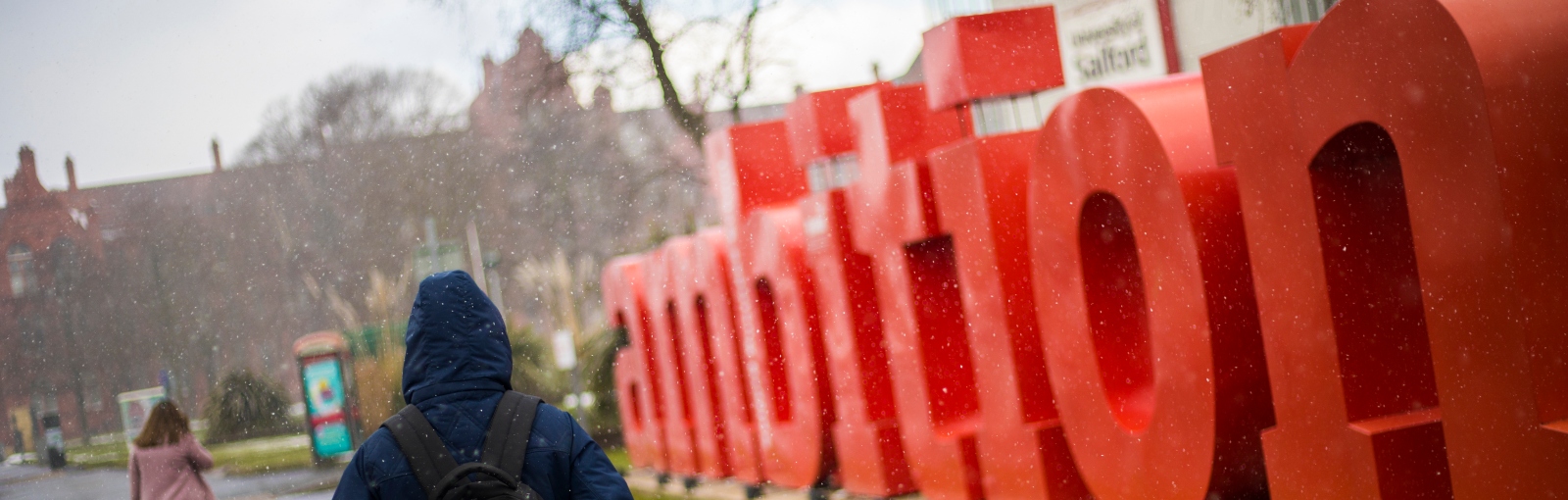 Photo of student wearing a blue jacket walking next to the ambition sign next to the Maxwell building