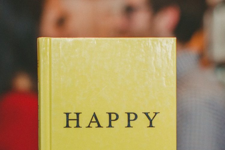 Photograph of a book with "Happy" written on the front cover, held up in front of a couple facing towards each other alongside some bookshelves