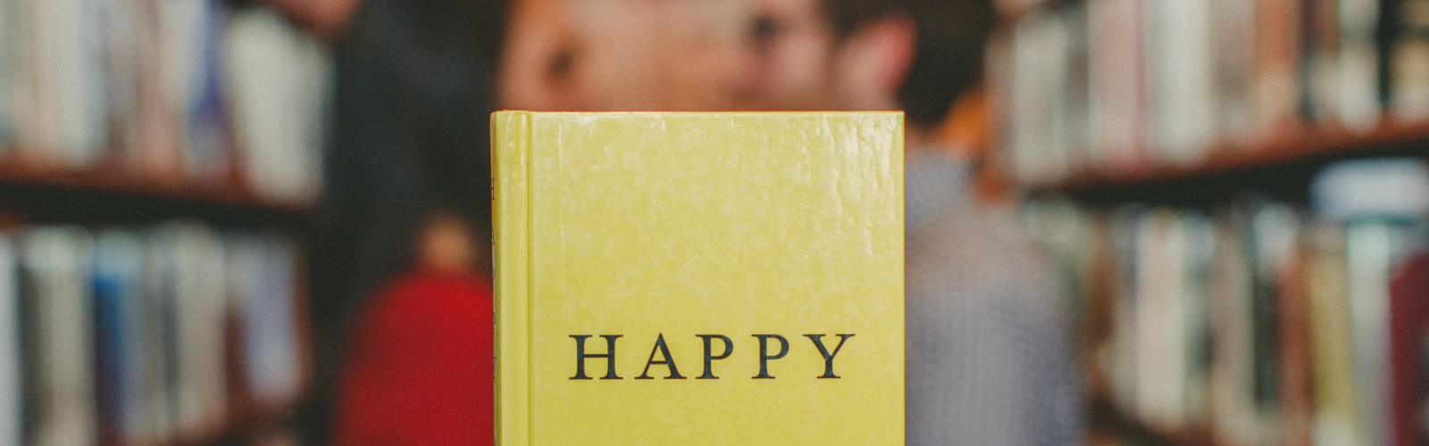 Photograph of a book with "Happy" written on the front cover, held up in front of a couple facing towards each other alongside some bookshelves