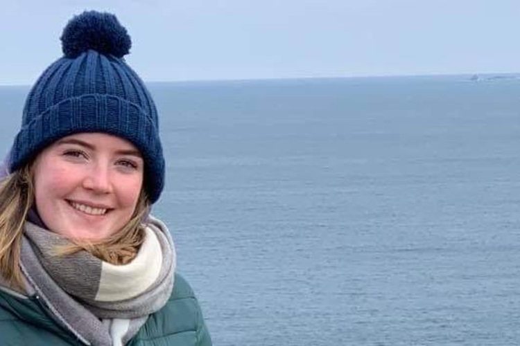 Niamh standing in front of sea. She is wearing a blue bobble hat, green coat and grey scarf.