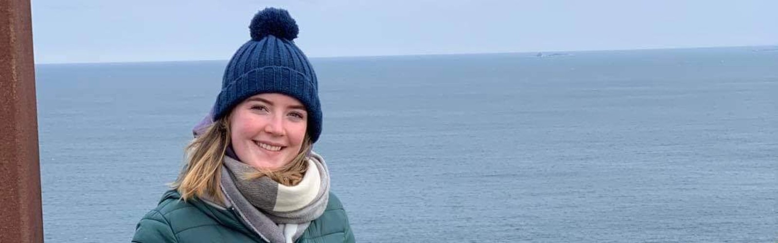 Niamh standing in front of sea. She is wearing a blue bobble hat, green coat and grey scarf.