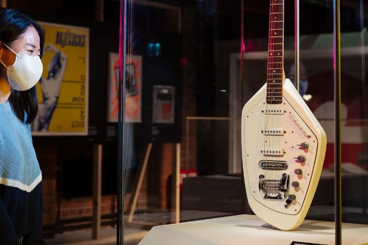 Photo of a women standing infront of a guitar that is placed is a glass box