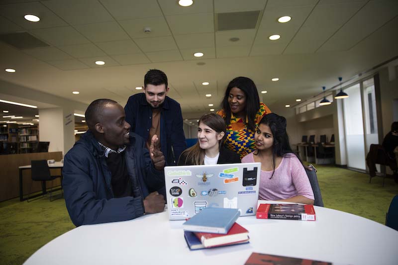 5 students around a table in the University of Salford's Clifford Whitworth library. There is a laptop and books on the table.