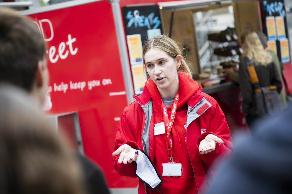 Photo of a student volunteer wearing red salford uni jacket and shirt talking to other students