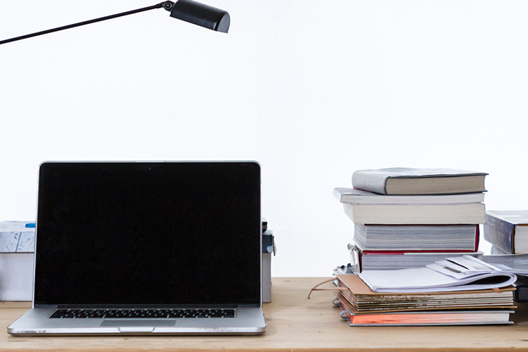 Photo of a latop, with desklamp, books, folder from let to right on top of wooden table