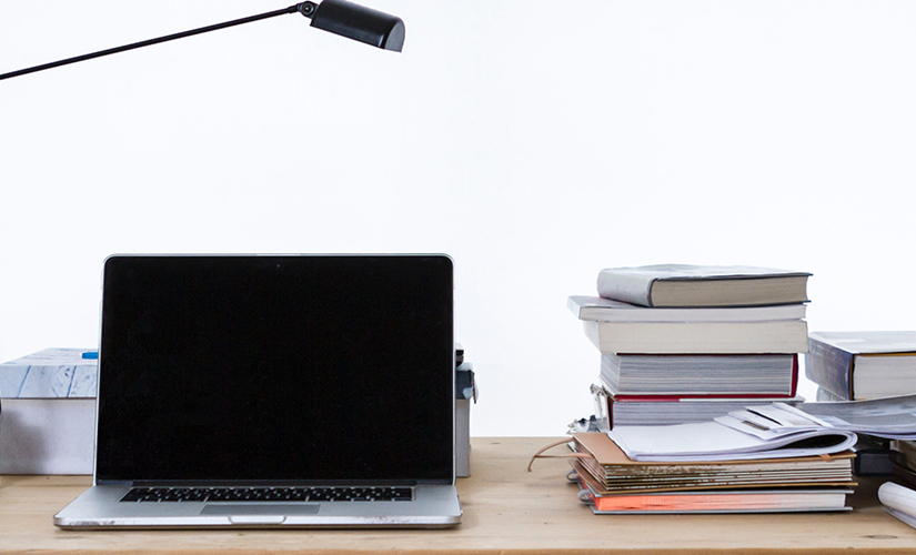 Photo of a latop, with desklamp, books, folder from let to right on top of wooden table