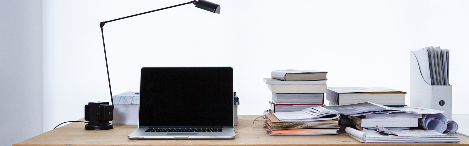 Photo of a latop, with desklamp, books, folder from let to right on top of wooden table