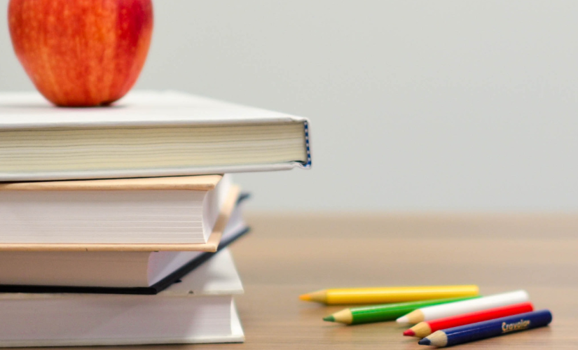 An apple placed on top of a stack of books with colored pencils and alphabet blocks to the right