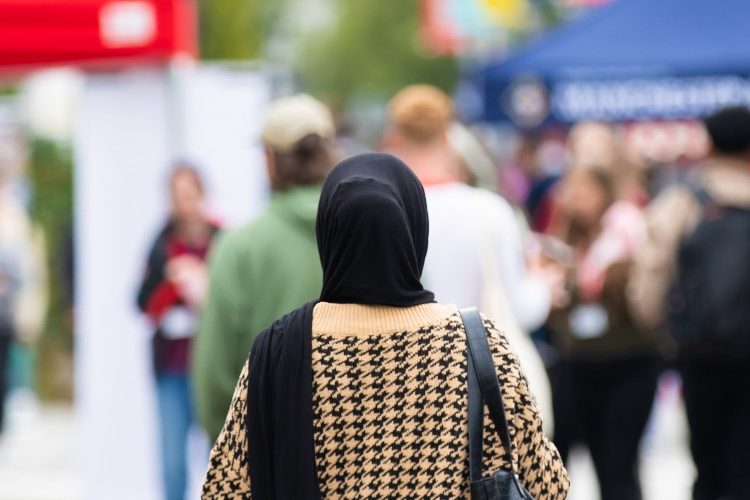 Photograph of Welcome Week at the University of Salford, outside, with a person in a headscarf in the centre of the frame, facing away from the camera. Stalls can be seen in the background.