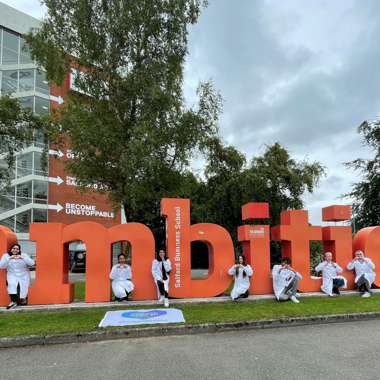 Students in lab coats post next to the 'ambition' sign outside of Salford's Maxwell building